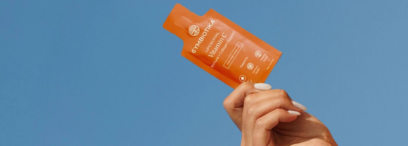 Hånd holder en pose med Cymbiotika Liposomal Vitamin C mot blå bakgrunn. Illustrasjonsbilde til artikkel om liposomal teknologi og kosttilskudd.